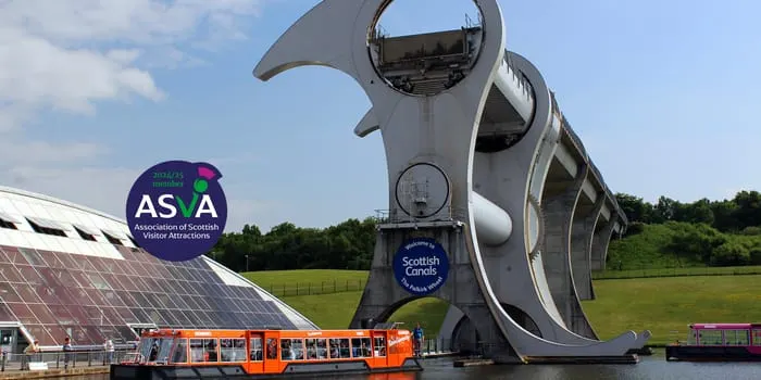 Orange tour boat passing beneath the Falkirk Wheel rotating boat lift structure over water, with solar panels and green fields visible.