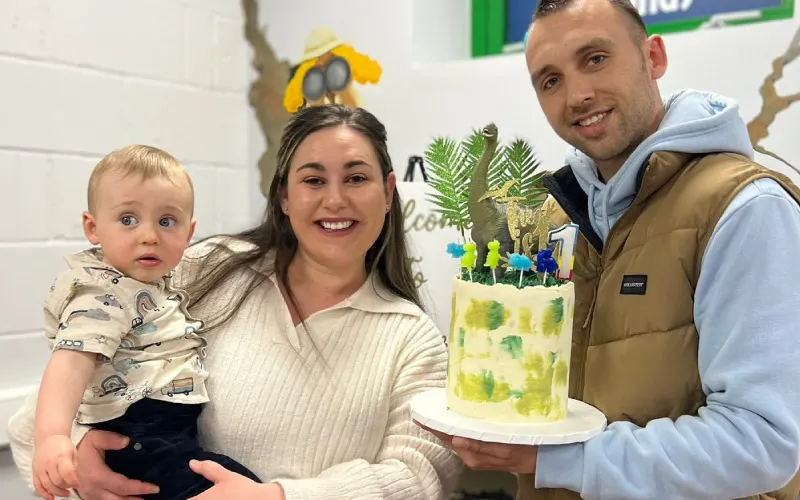 A woman holding a toddler stands with a man displaying a decorated cake with toy animals and plants.