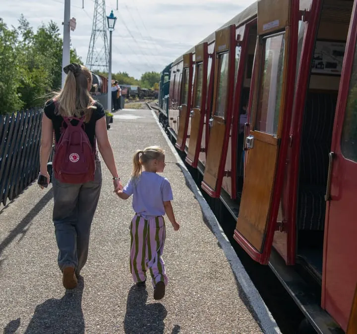 Adult and child walking hand-in-hand alongside vintage railway carriages at a heritage railway station.