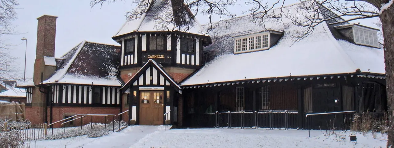 Tudor-style brick building with black and white timber framing covered in snow, entrance visible with bare trees overhead.