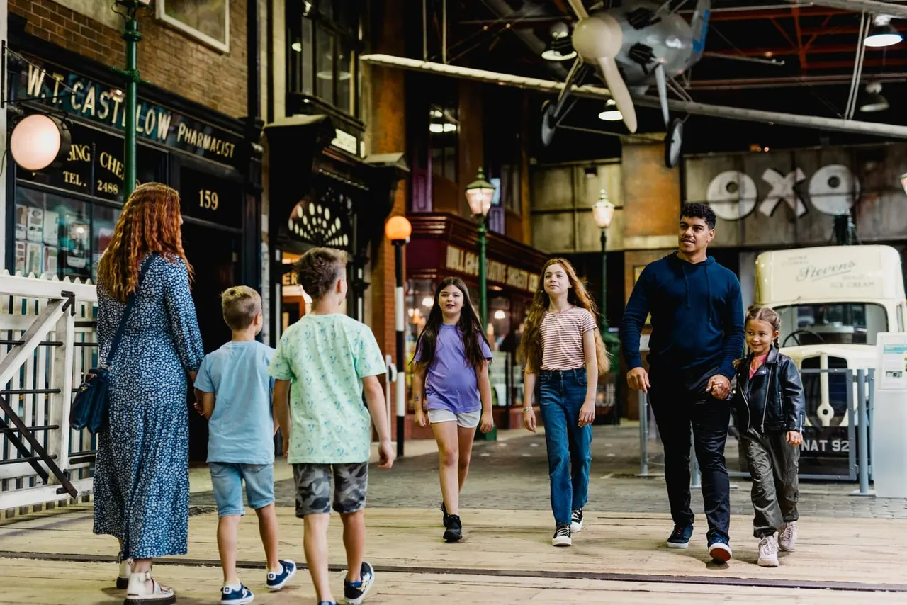 Family walking through a recreated historic street scene at night with period shopfronts and vintage vehicles.
