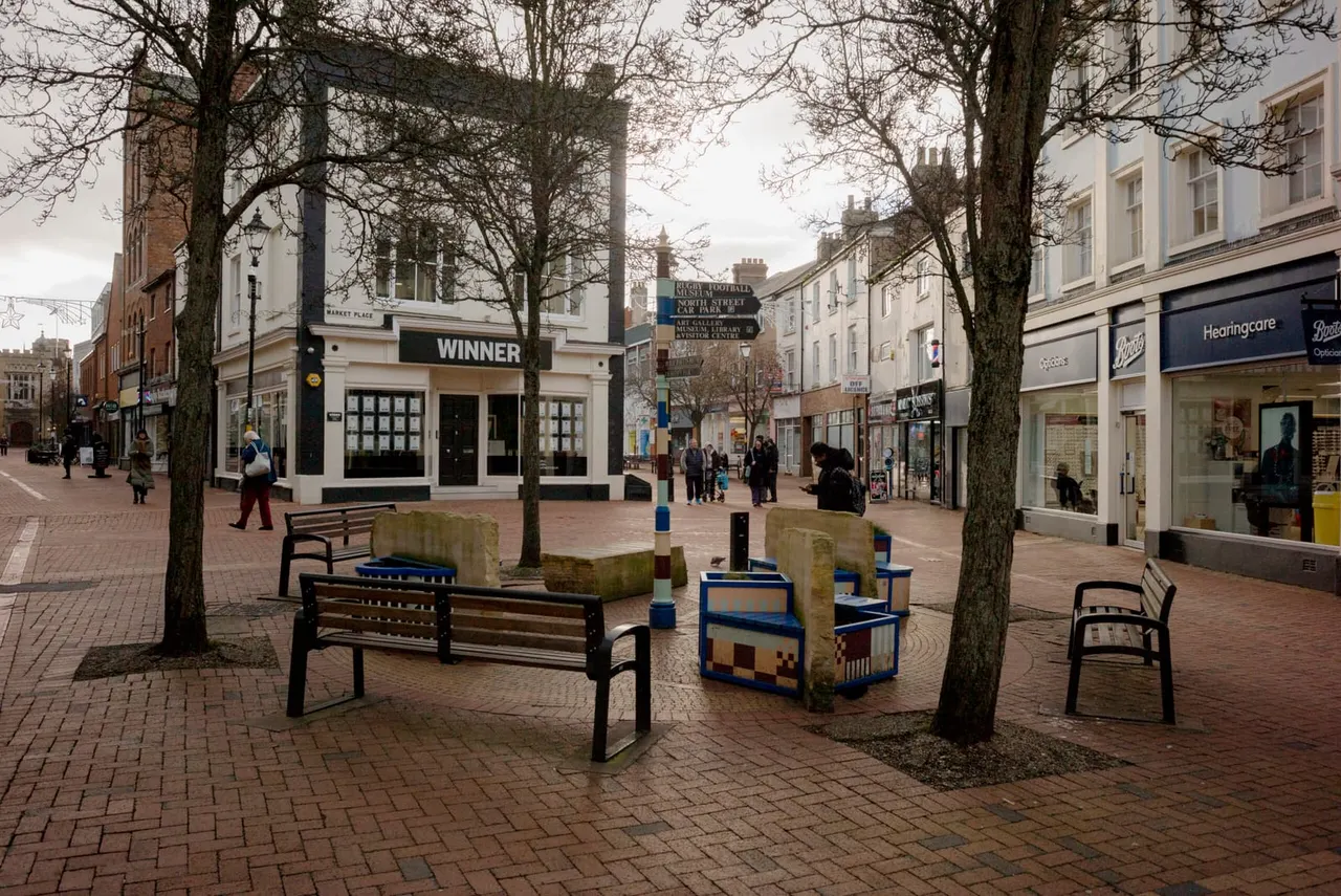 Town centre pedestrian street with benches, bare trees, shops and buildings, a few people walking.