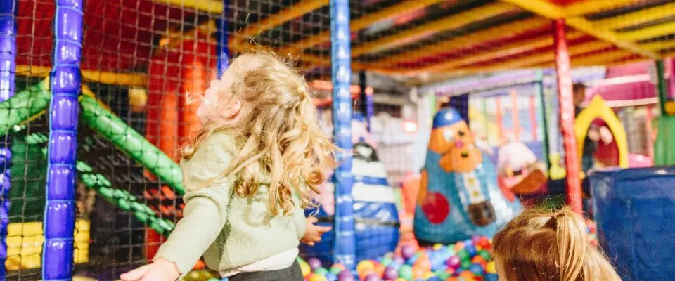 Young children playing in a colourful indoor soft play area with ball pit and climbing equipment.