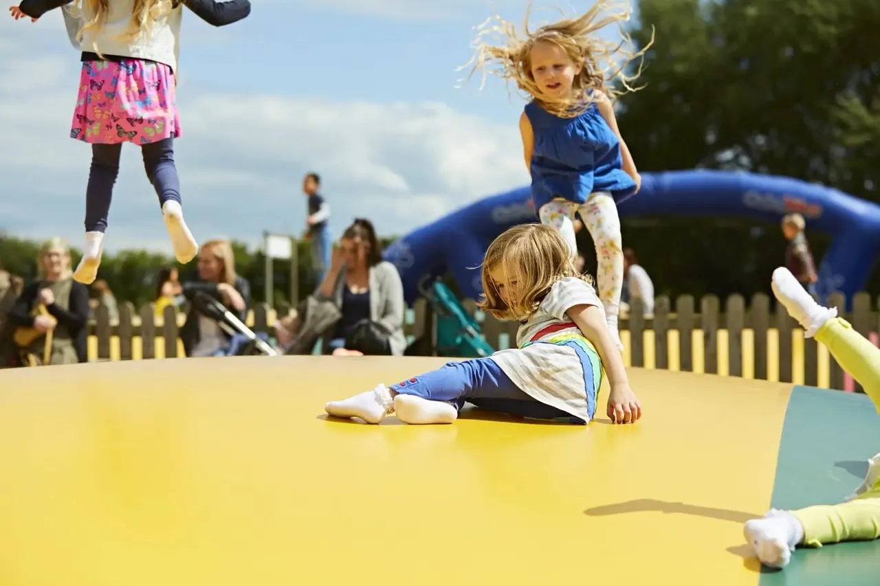 Young children jumping and playing on a large yellow inflatable trampoline with spectators watching nearby.