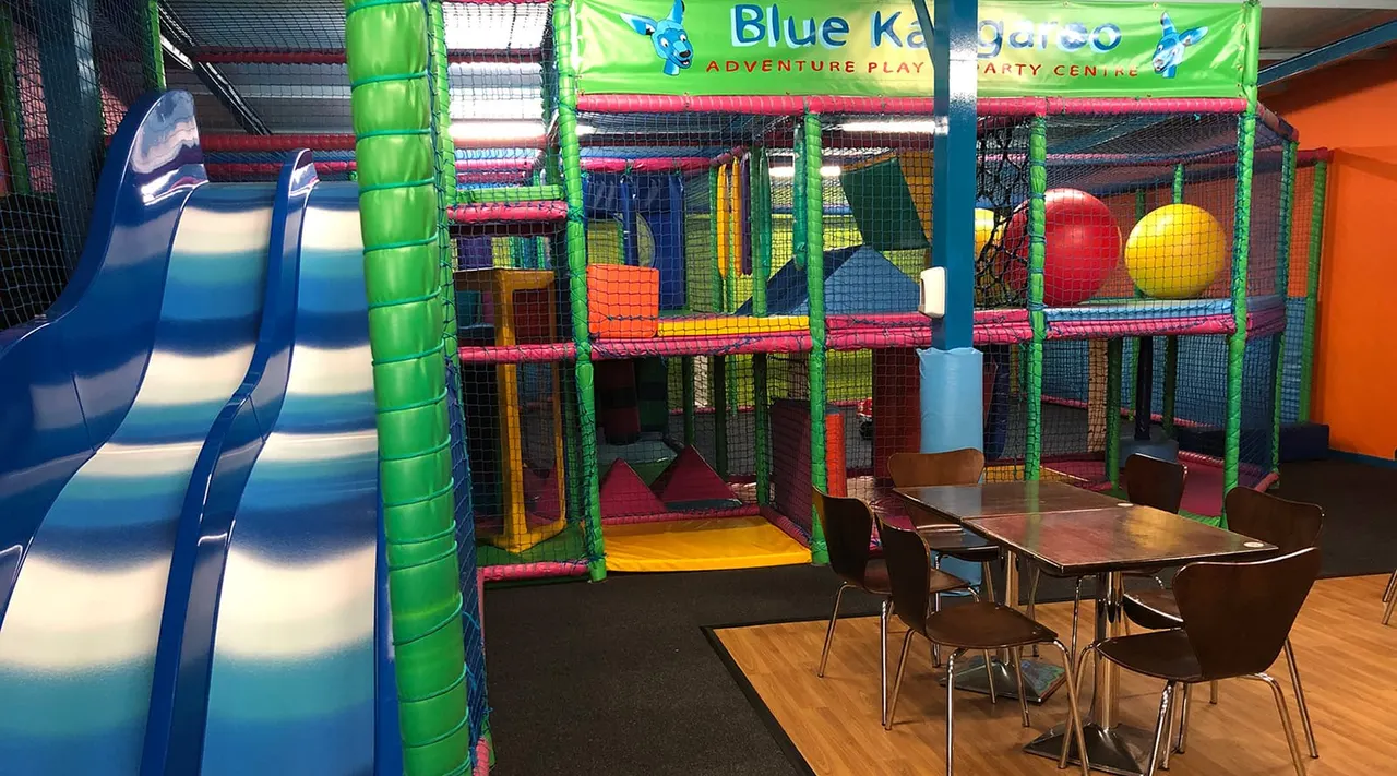 Colourful indoor play structure with slide, climbing frame and ball pit, seating area with tables and chairs in foreground.