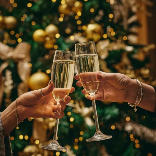 Two people clinking champagne glasses in front of a decorated Christmas tree with gold baubles.