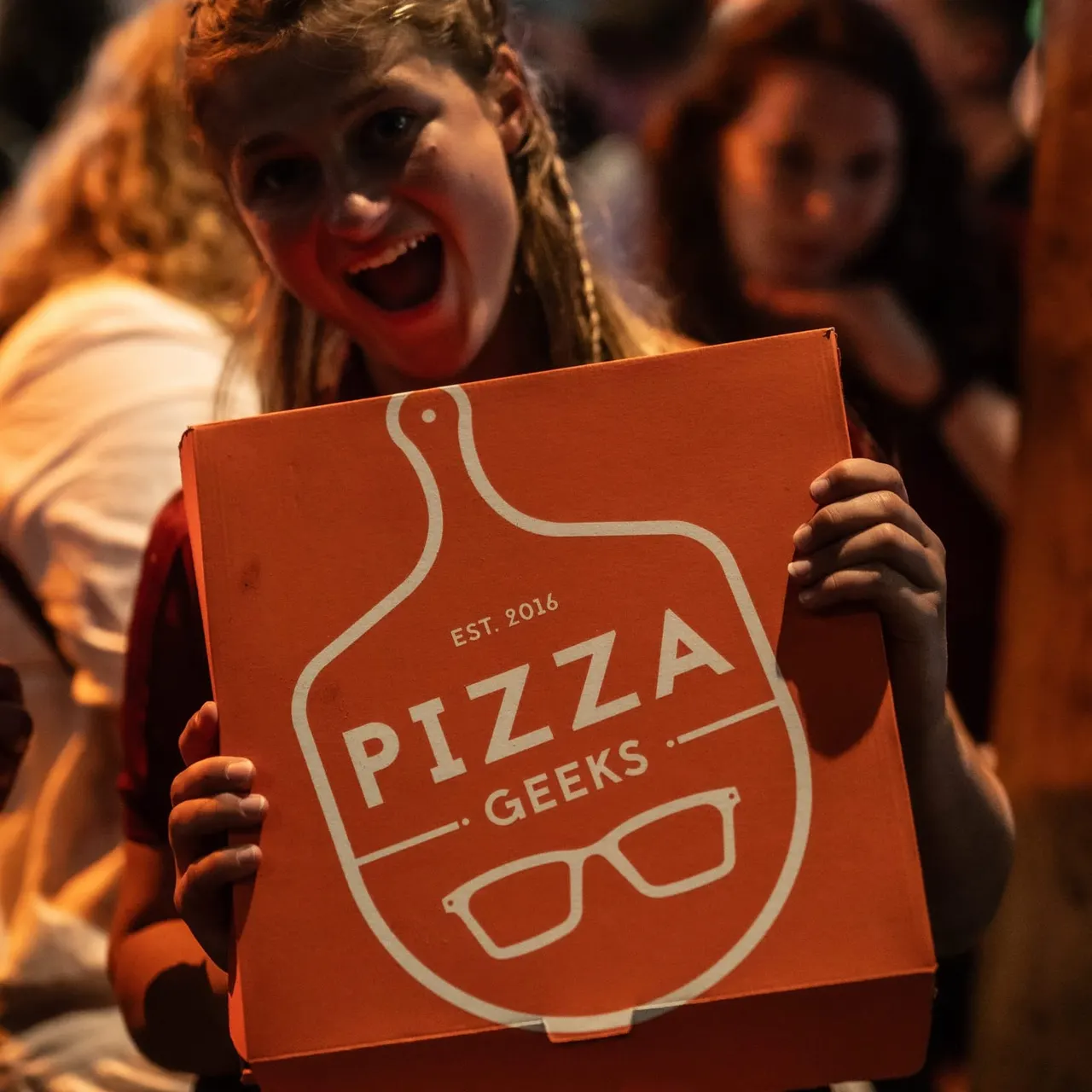 Child holding an orange Pizza Geeks box whilst smiling at an indoor event with other people visible in the background.
