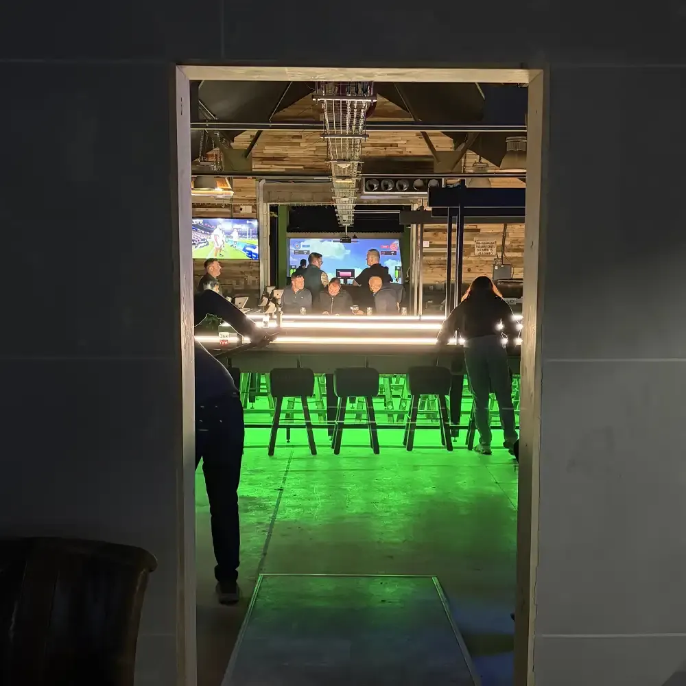 Bar interior with neon-lit counter, green barstool seating, patrons and staff, and screens showing sports.