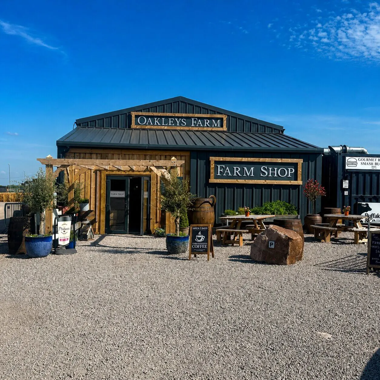 Modern farm shop building with dark metal roof and wooden pergola entrance, set on gravel with plants and rustic décor under blue sky.