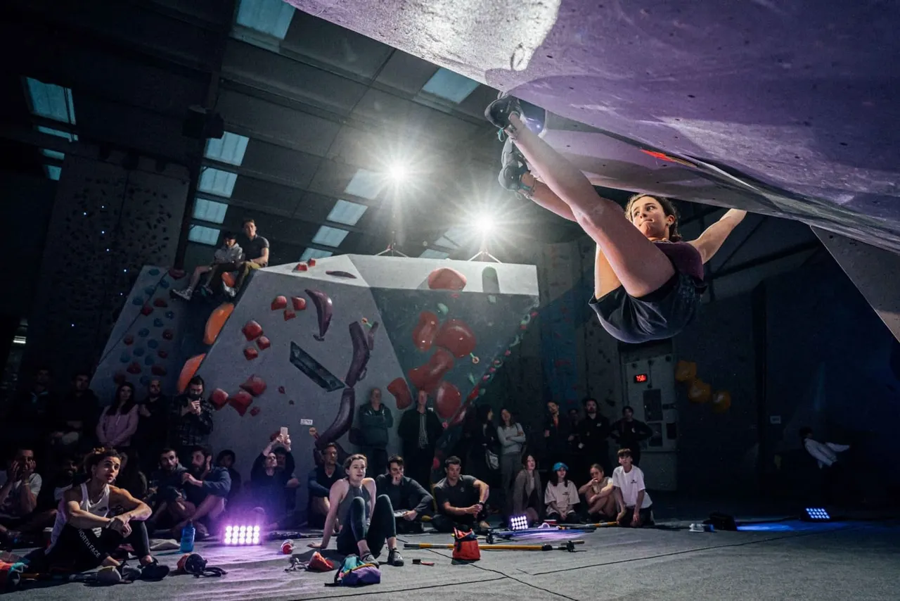 Person indoor rock climbing on an overhanging wall whilst spectators watch from ground level.