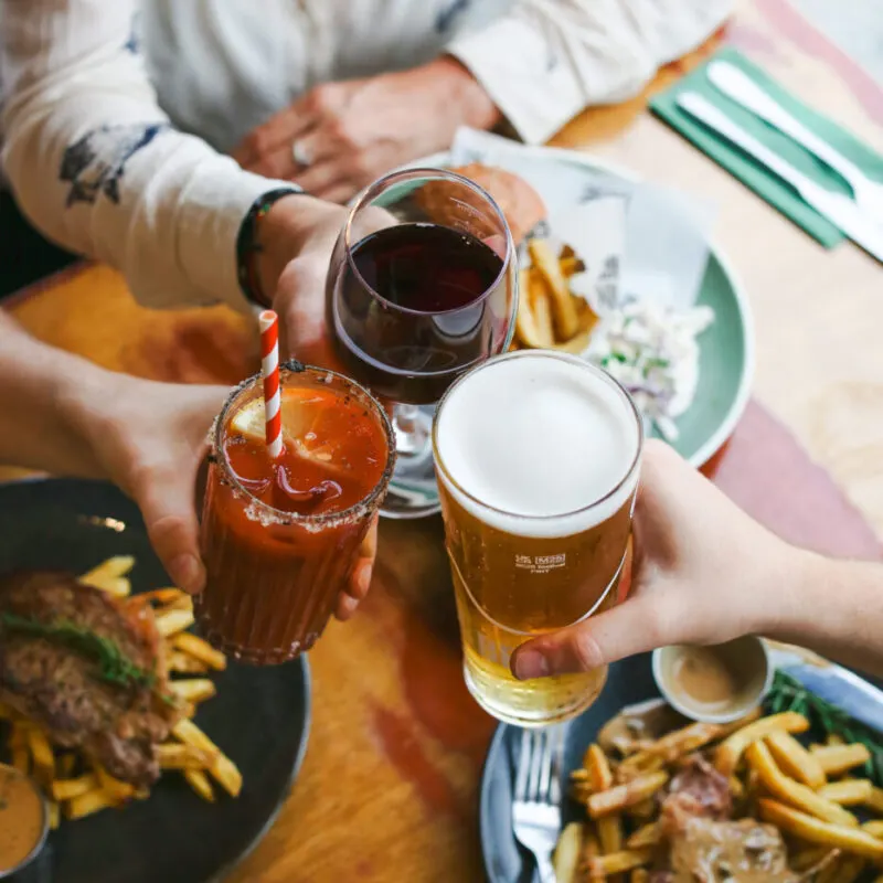 Two people toasting with beer and soft drinks at a dining table with food and fries.