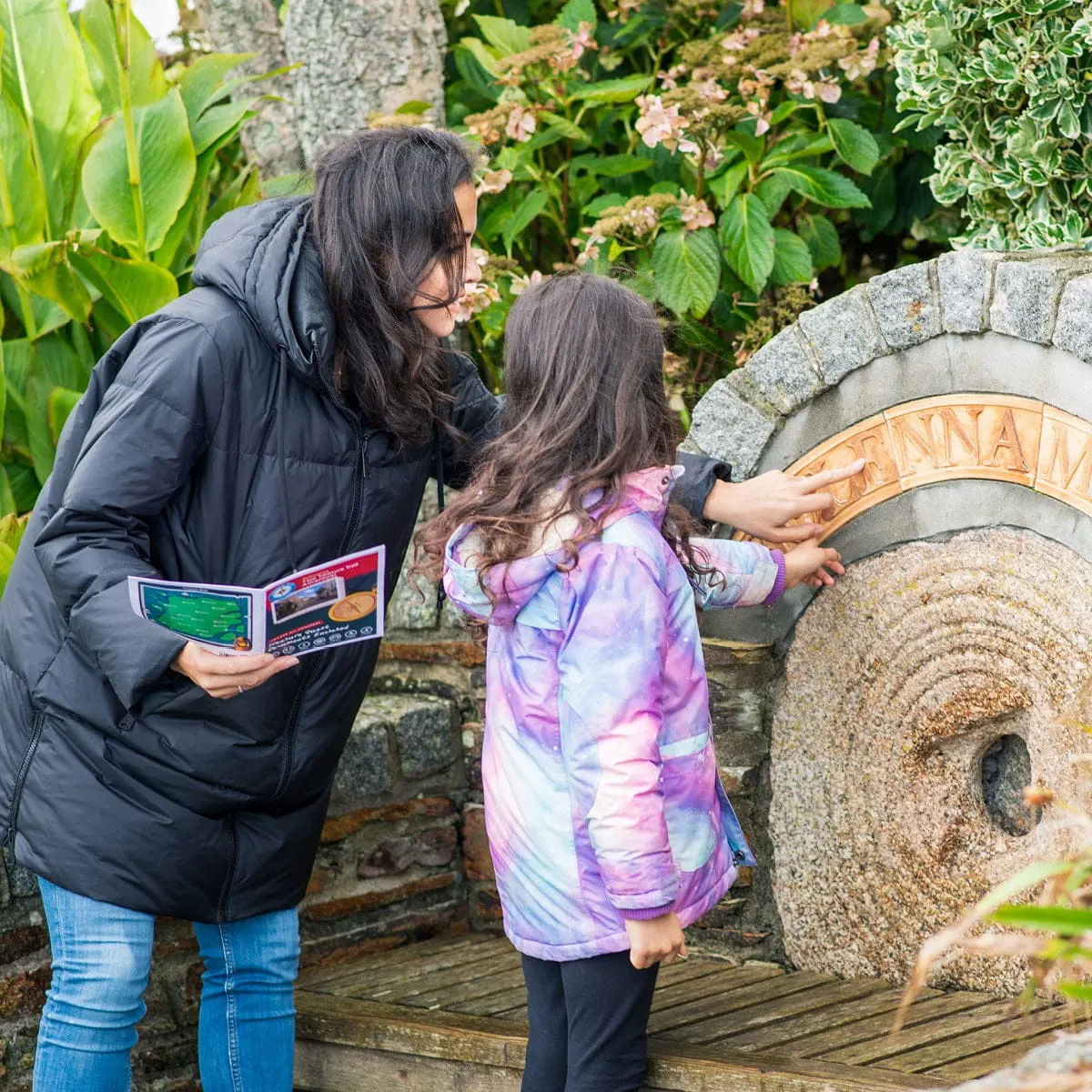 An adult and child examine a stone millwheel set amongst flowering plants and greenery on a garden trail.