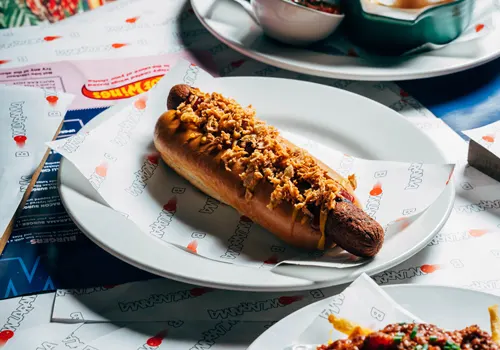 A hot dog topped with crushed peanuts sits on a white plate at a dining table.