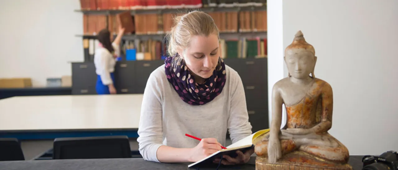 Woman writing in notebook at desk with Buddha statue beside her and bookshelves in background.