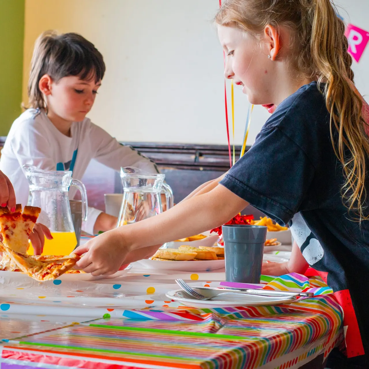 Two children at a table with colourful decorations, water jugs and plates of food during a party or celebration event.