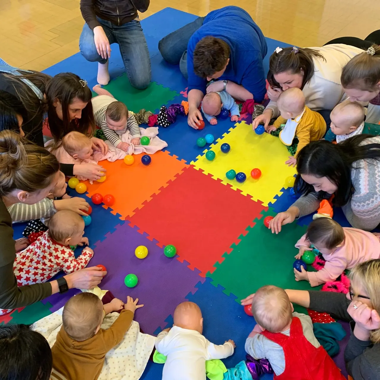 Adults and babies gathered around a colourful foam play mat with balls, engaging in sensory play together.