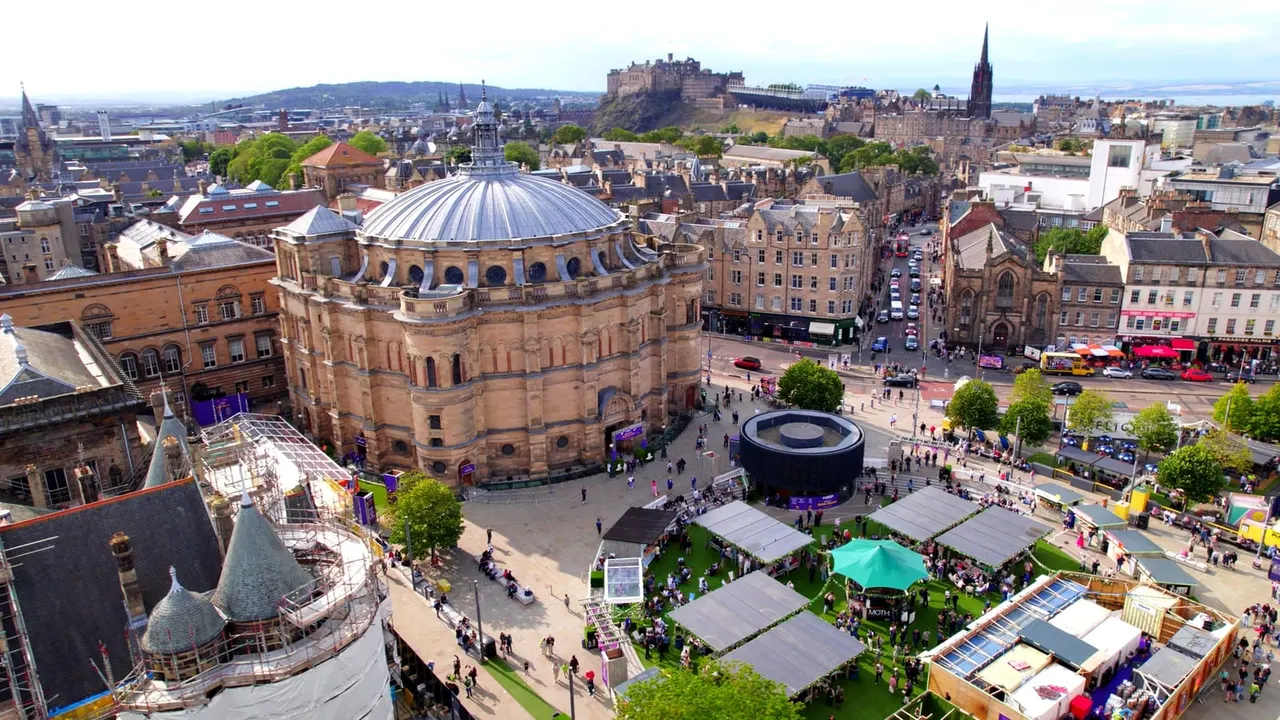 Aerial view of Edinburgh city centre with a domed historic building, castle on the skyline, and a crowded public square with market stalls and umbrellas below.