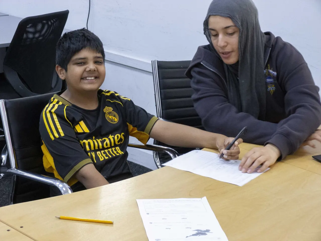 Young boy in sports shirt signing a document whilst smiling, with a staff member assisting at a table.
