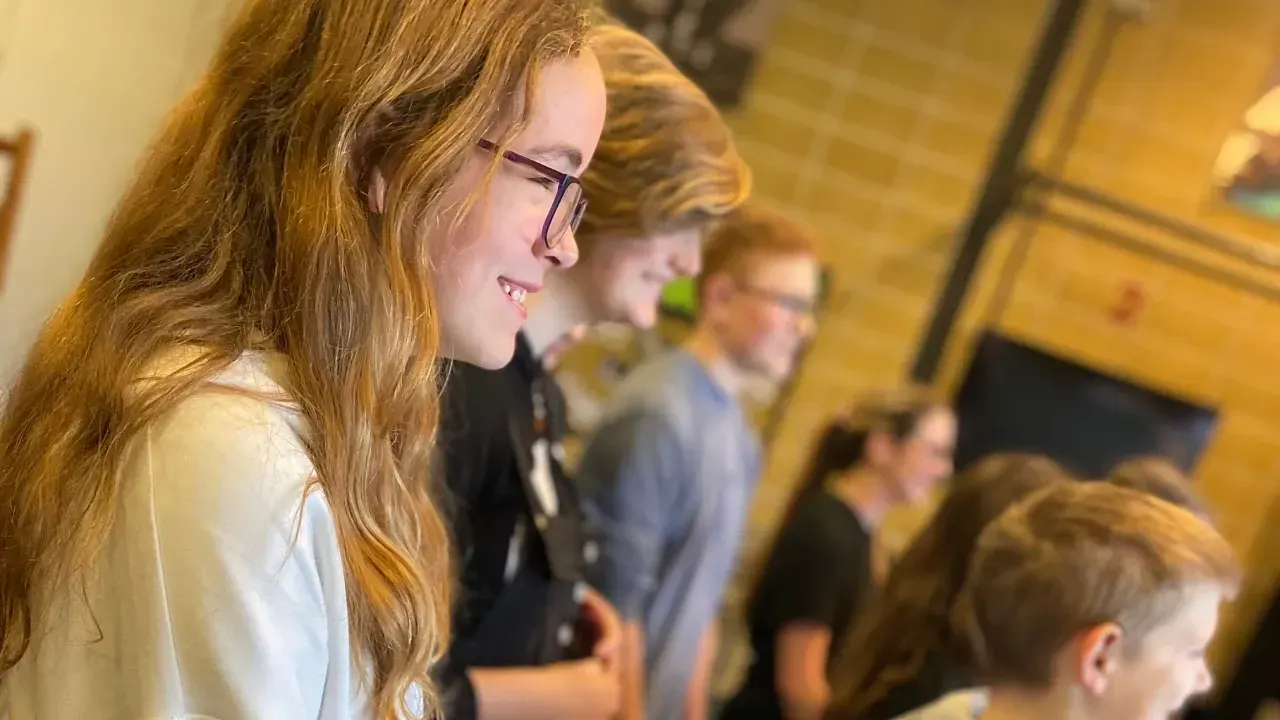 A young woman wearing glasses smiles whilst standing indoors with other visitors in the background.
