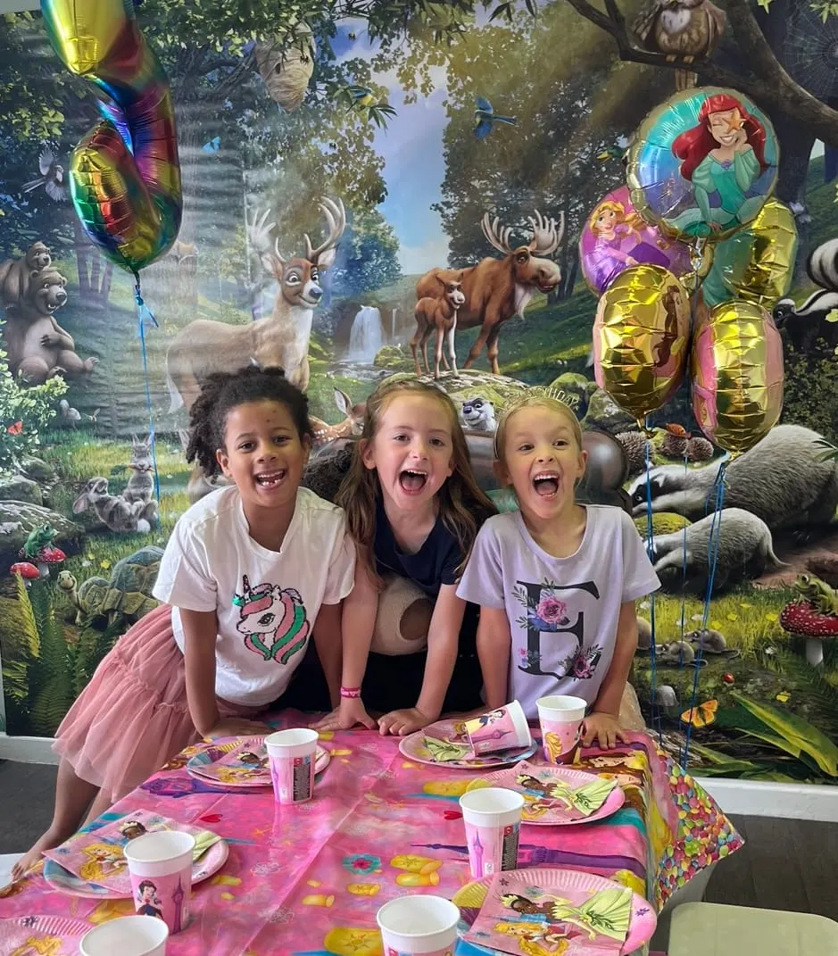 Three children at a colourful party table with themed decorations, balloons and fantasy forest backdrop.