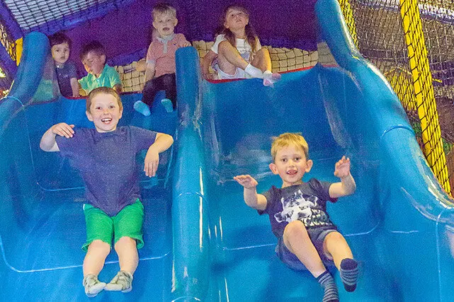 Children sliding down blue slides in an indoor soft play area.