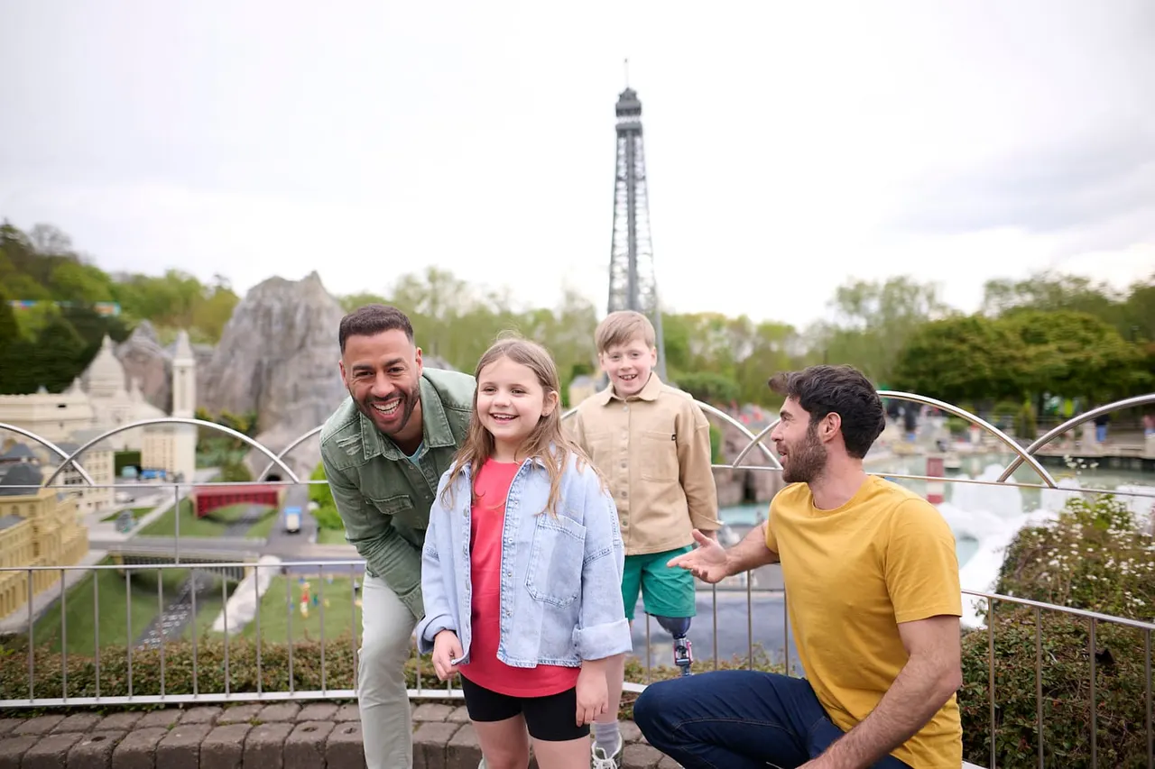 Two adults and two children smiling at a viewing point overlooking a miniature fairground with a tall tower structure in the background.