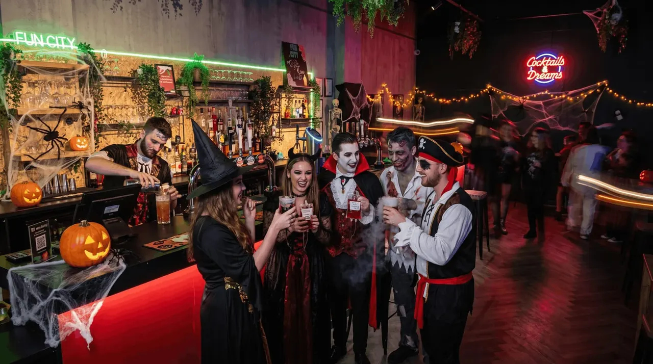 People in Halloween costumes enjoying drinks at a themed bar with neon signs, jack-o'-lanterns and spooky decorations.
