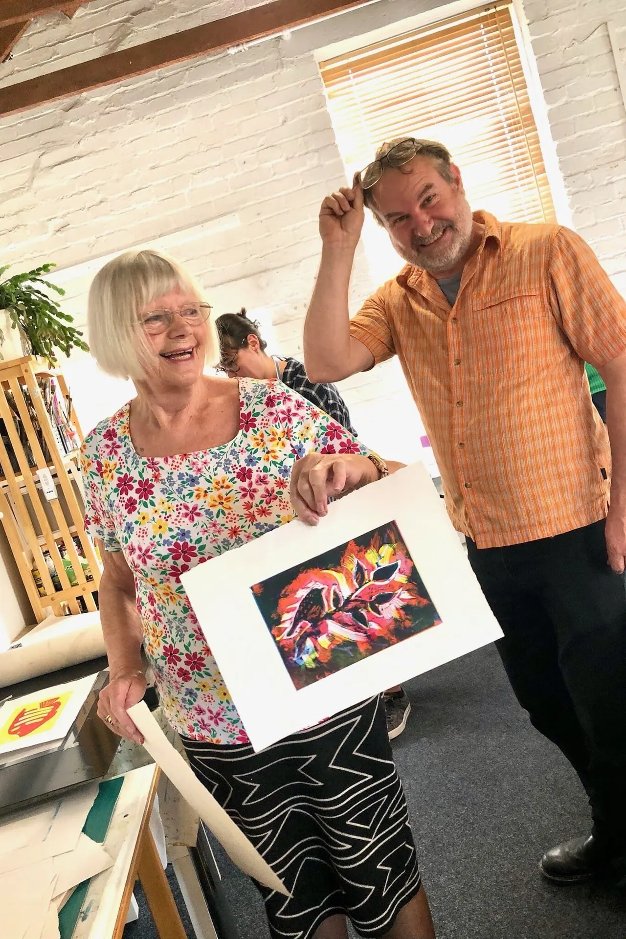 Two adults displaying a colourful floral artwork in an indoor studio space with white brick walls and natural light.