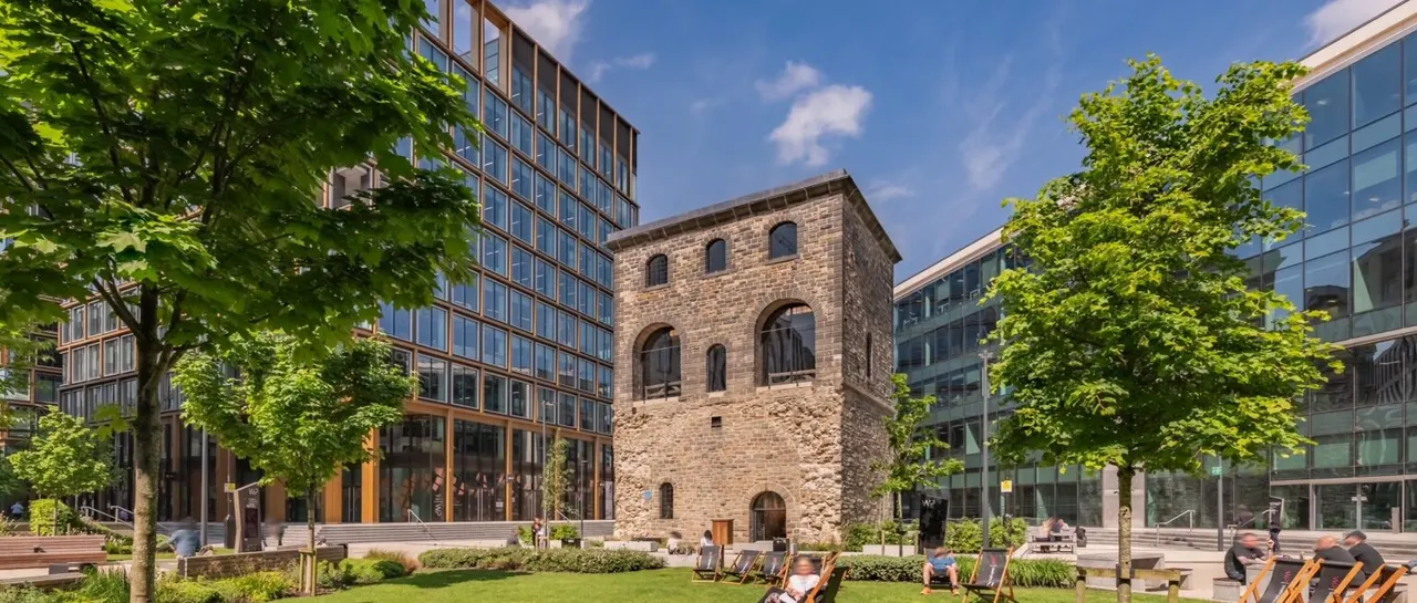 Urban courtyard with visitors relaxing on deckchairs beneath green trees, surrounded by modern glass buildings and a historic stone tower.