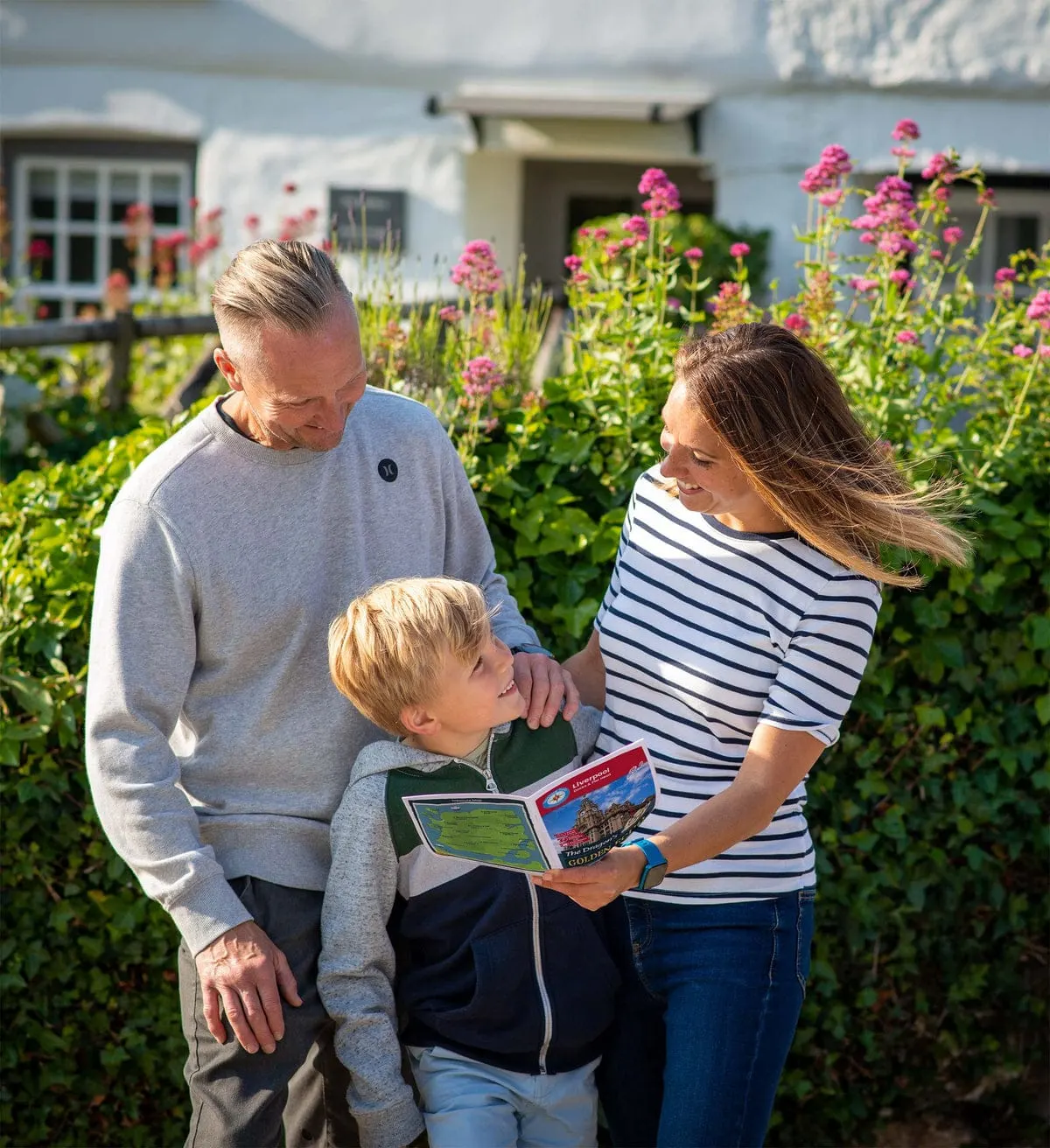 Family with young child looking at a map in a flower garden with pink blooms and holiday accommodation buildings behind.