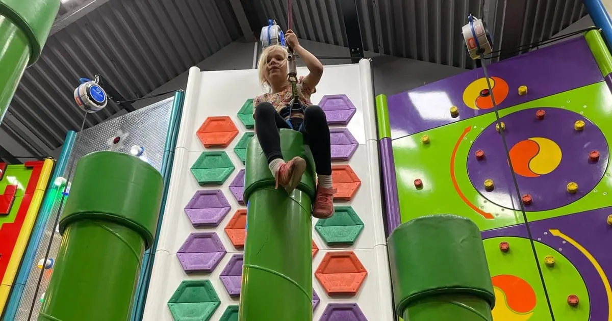 A child climbing an indoor climbing wall with coloured hexagonal holds, reaching upward with climbing equipment.