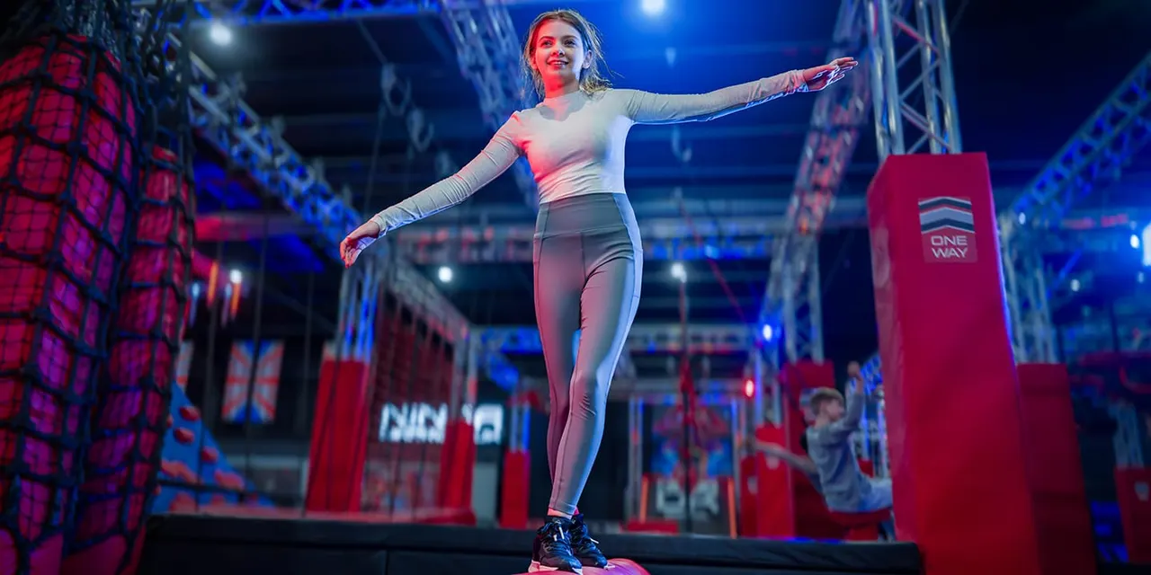 Woman balancing on an obstacle course platform with arms outstretched in a neon-lit indoor arena.