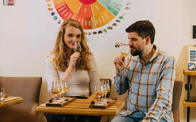 A man and woman sit together tasting drinks from glasses on a wooden table beneath a colourful wheel chart on the wall.