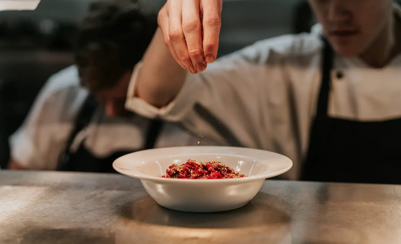 A chef sprinkling red seasoning onto a dish in a white bowl during a cooking demonstration.