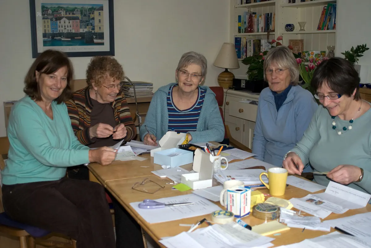 Five women seated around a wooden table with papers and refreshments during an indoor activity or meeting.
