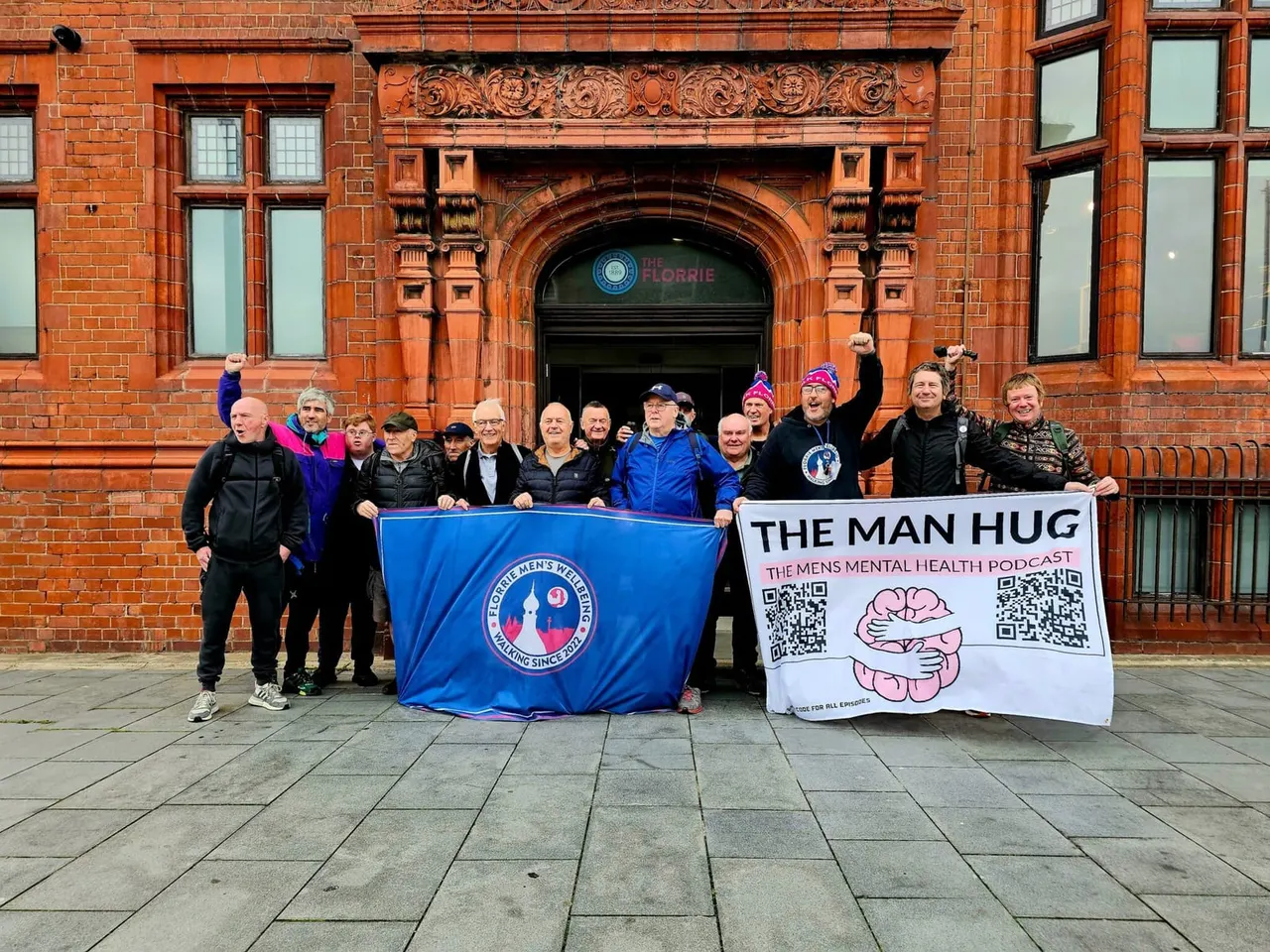 A group of people stand outside a red brick building holding banners for a men's mental health podcast and organisation.