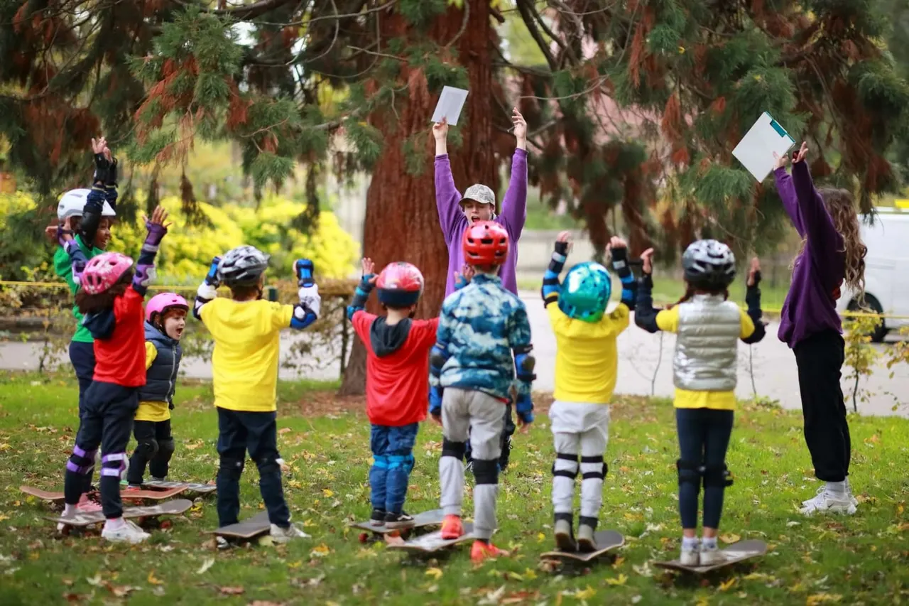 Children in skateboarding protective gear celebrating with raised arms and certificates beneath tall conifer trees in a park.