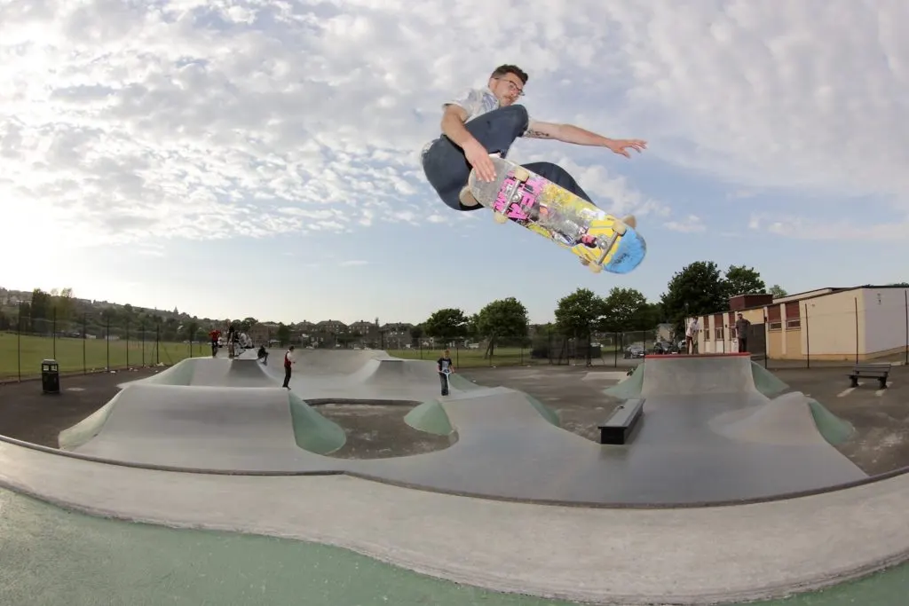 A skateboarder performs a trick mid-air above concrete ramps at an outdoor skate park.