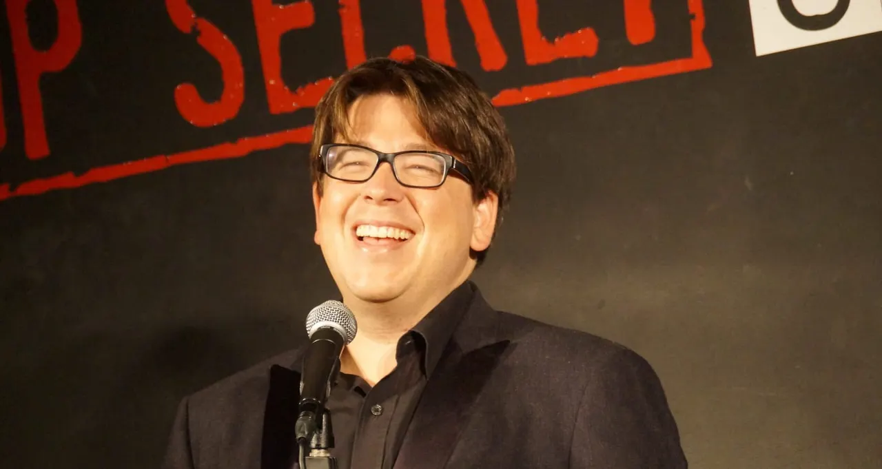 Man wearing glasses speaking at a microphone on stage at a comedy club.