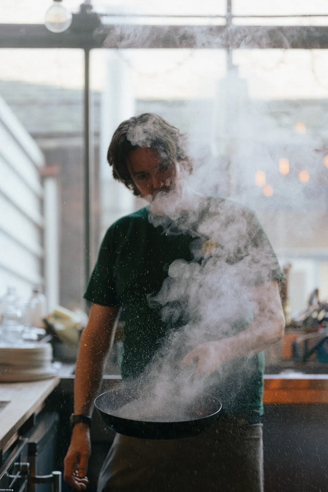 Chef tossing flour or powder from a pan in a kitchen with city views through windows behind.