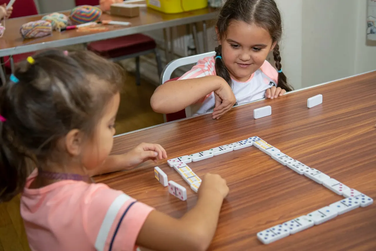 Two young children playing dominoes at a wooden table during an indoor activity session.