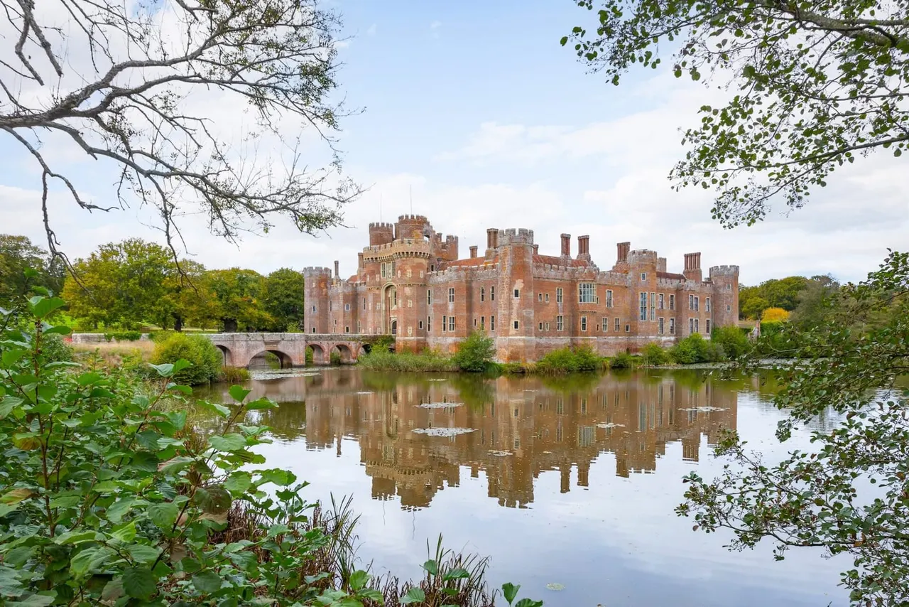 Red brick castle with crenellated towers reflected in still water, surrounded by trees and vegetation.