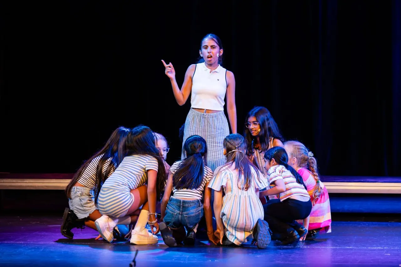 Young woman performing on stage with a group of children gathered around her under blue lighting.