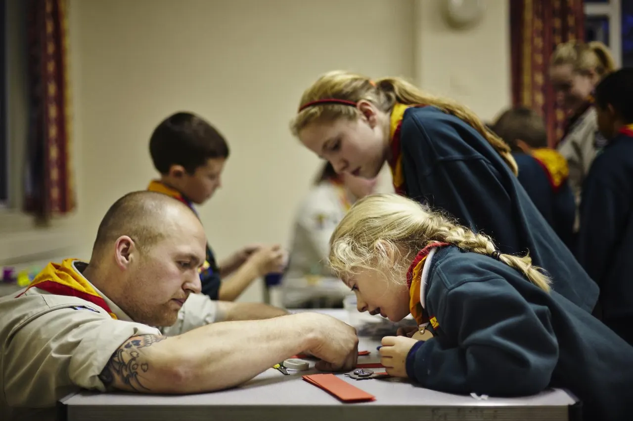 An adult scout leader helps young children with a craft or badge activity at a table indoors.