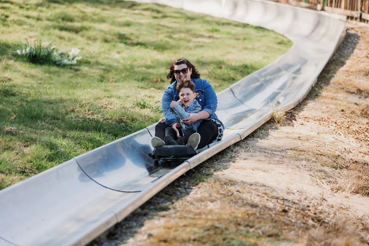 Adult and young child sledging down a twisting slide at an outdoor leisure park.