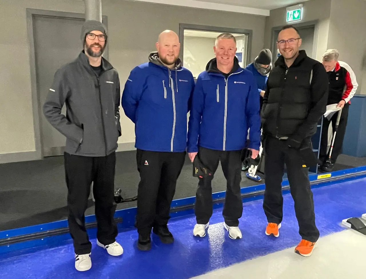 Group of six people standing on an indoor curling rink, dressed in winter jackets and trainers.
