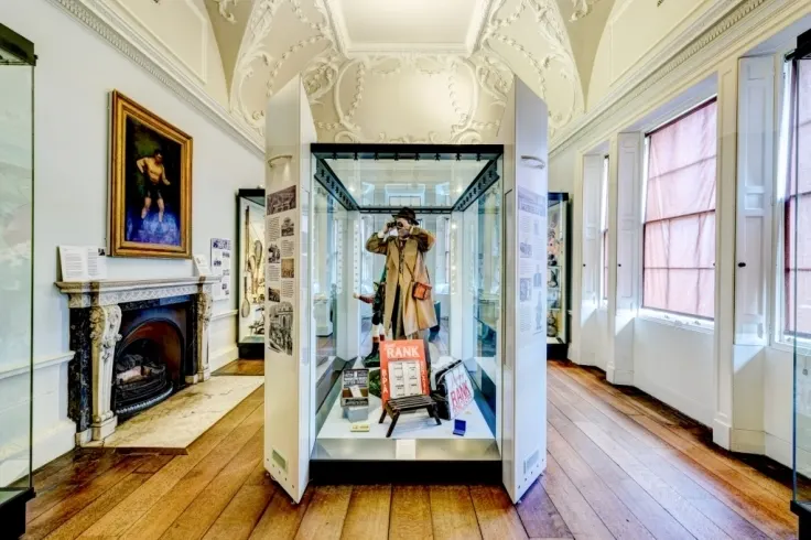 Historic museum room with ornate plasterwork ceiling, fireplace, portrait painting, and central display case showing period clothing and accessories.