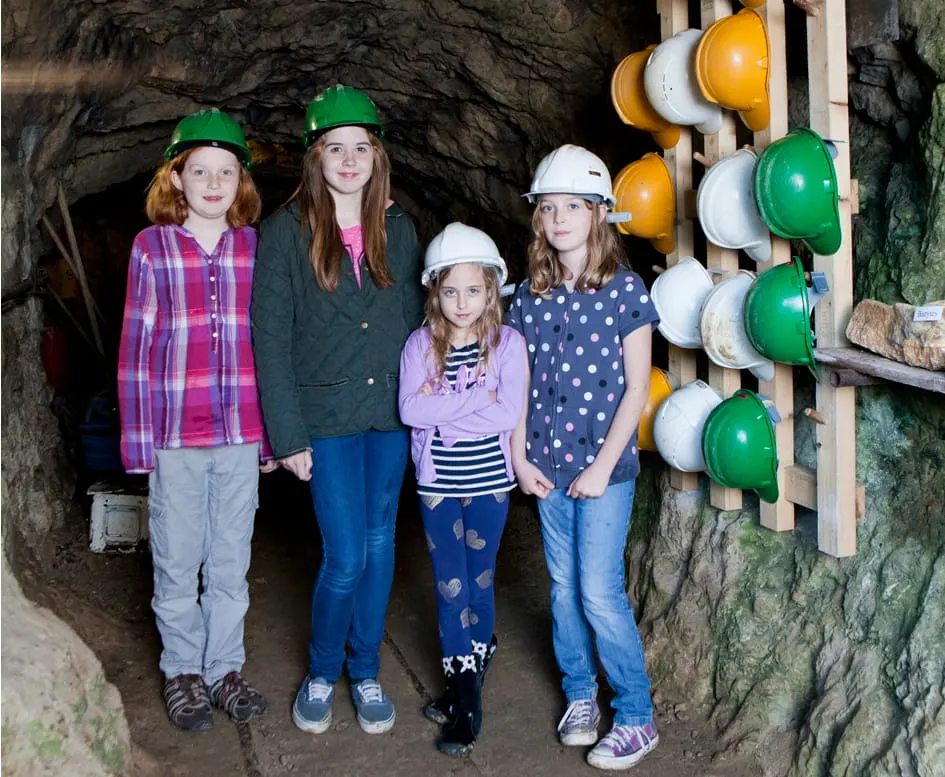 Four children wearing safety helmets stand in a mine tunnel next to a rack of coloured hard hats.