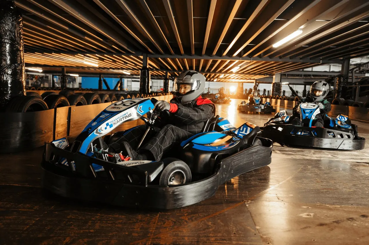 Two drivers in helmets race blue and black go-karts on an indoor track with wooden barriers and spectators watching.