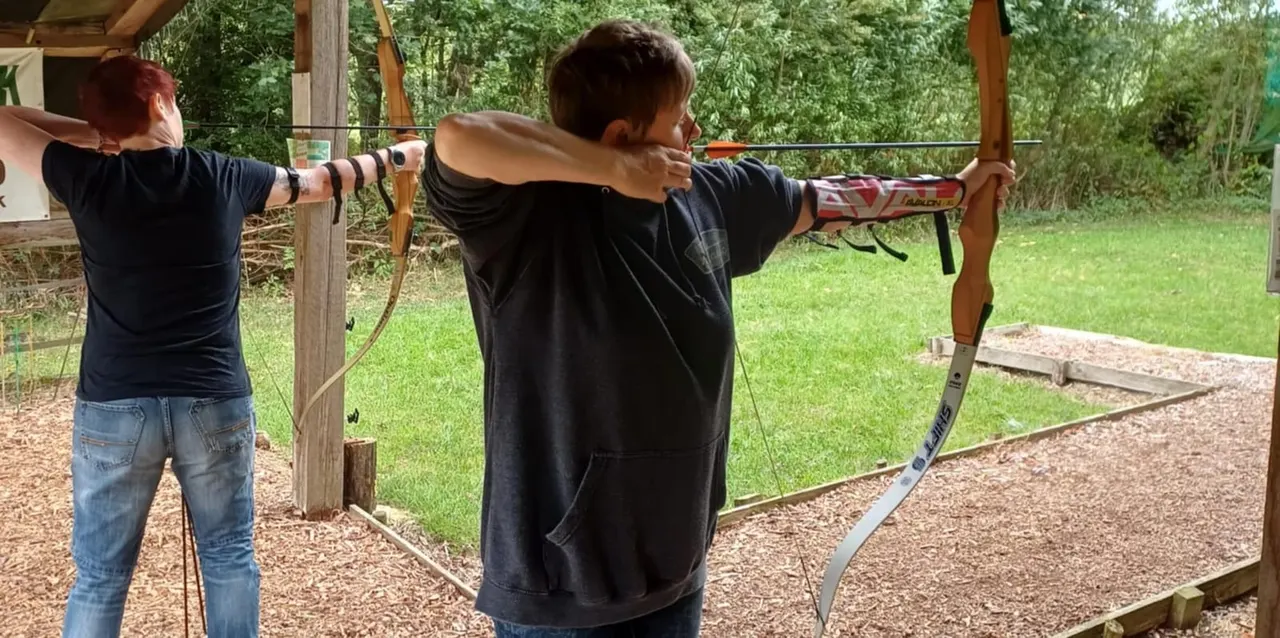 Two people practising archery outdoors, drawing wooden bows at a range with woodland backdrop.